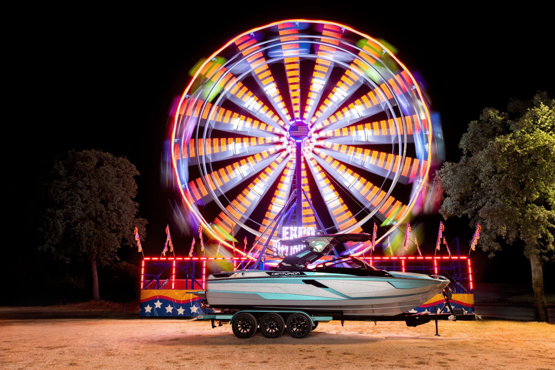 A brightly lit Ferris wheel spins at night behind a parked 2026 boat Ri245 on a trailer, with trees on either side and dry grass in the foreground—hinting at water sports adventures to come.
