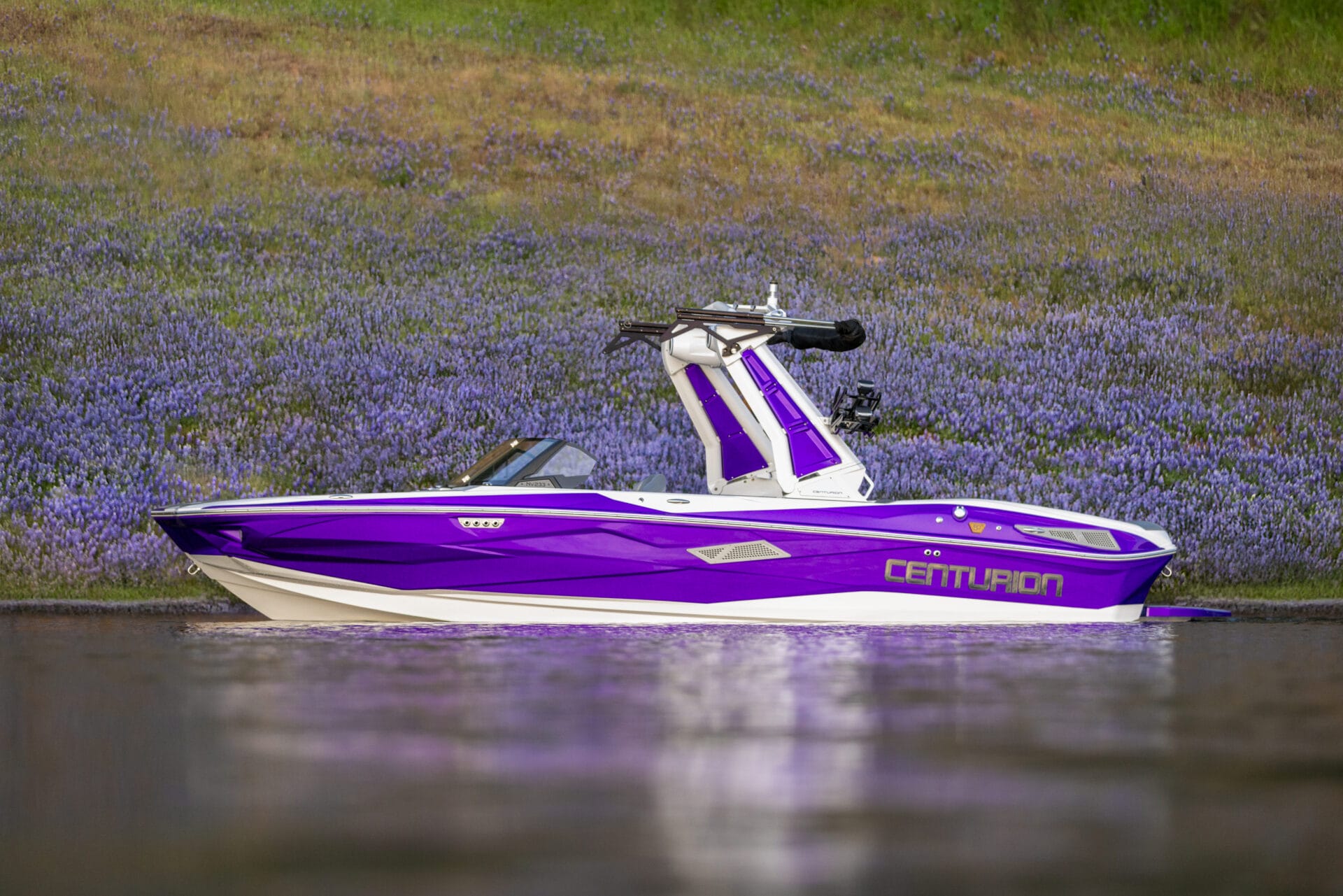 A purple and white Centurion speedboat is stationary on calm water, with a grassy, purple-flowered hillside in the background.