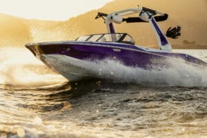 A purple and white speedboat moves quickly on the water, creating splashes, with the sun setting over hills in the background.