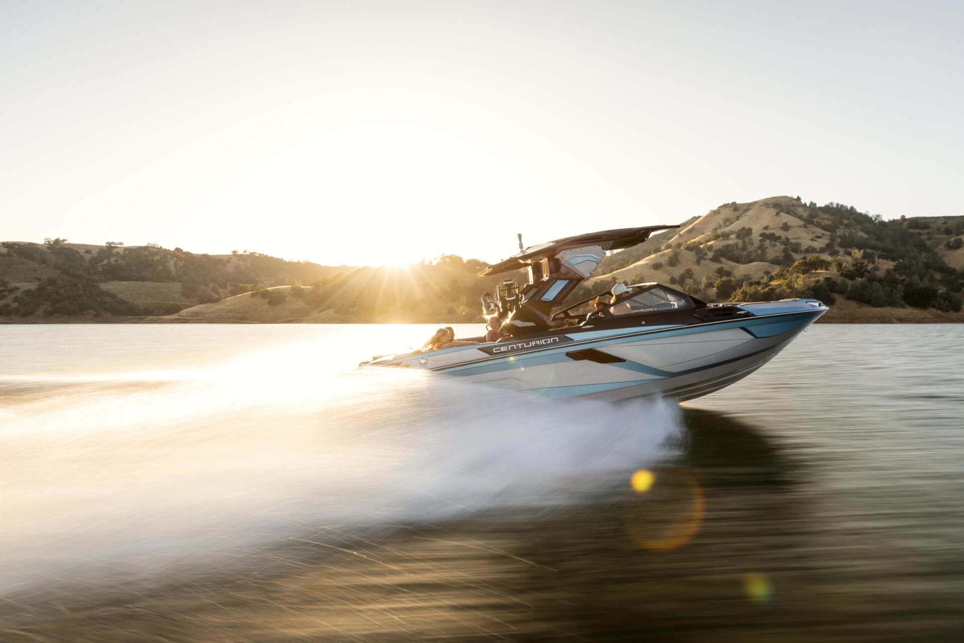 A speedboat moves quickly across a lake at sunset, with hills and trees visible in the background.