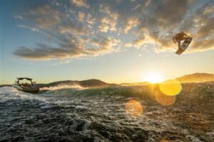 A person wakeboarding is airborne behind a motorboat on a lake at sunset, with water splashing and sun flares visible.