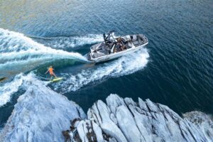 A person wakeboards behind a speedboat on a lake near rocky shoreline, with several passengers visible in the boat.