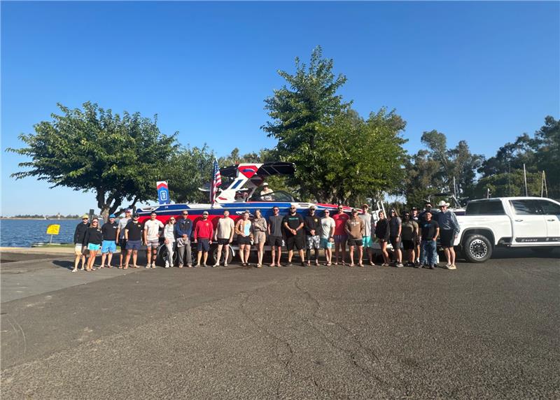 A group of people stands in front of a boat hitched to a white truck at a lakeside parking area, with trees and water visible in the background.