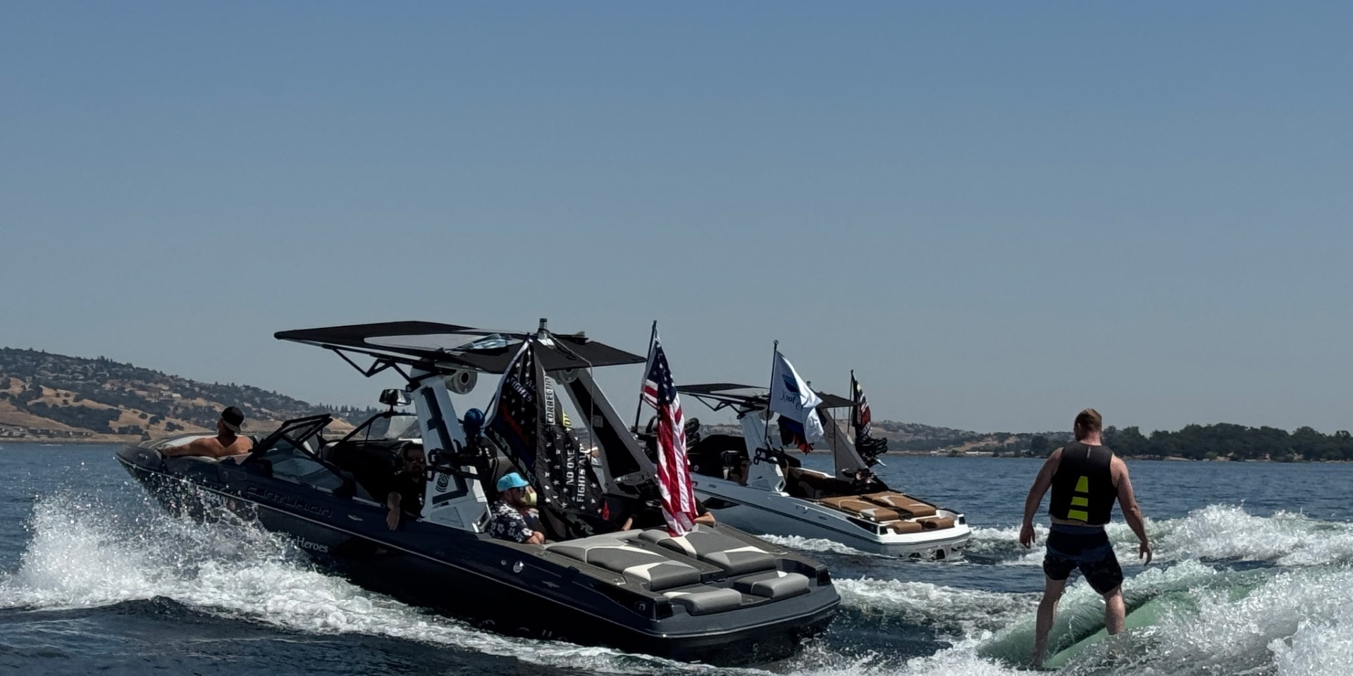 Two boats with flags tow people on a lake, while a man in a life vest wakesurfs behind one of the boats under a clear sky.