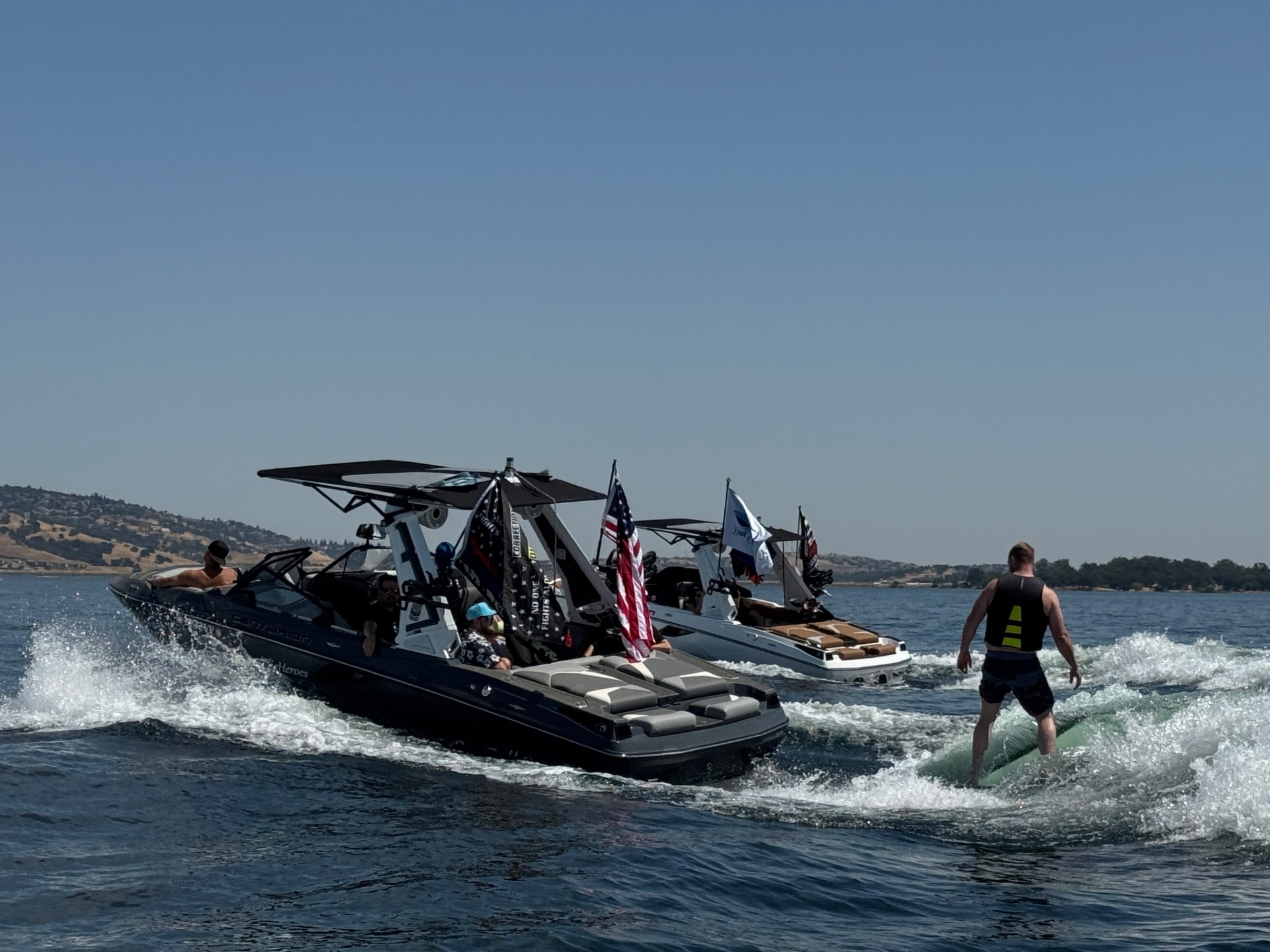 Two boats with flags tow people on a lake, while a man in a life vest wakesurfs behind one of the boats under a clear sky.