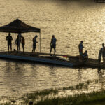A group of people stand and sit on a floating dock with a canopy at sunset, next to a calm body of water.