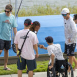 A group of people, including a person in a wheelchair, gather outdoors near a blue platform by the water; two people are shaking hands.