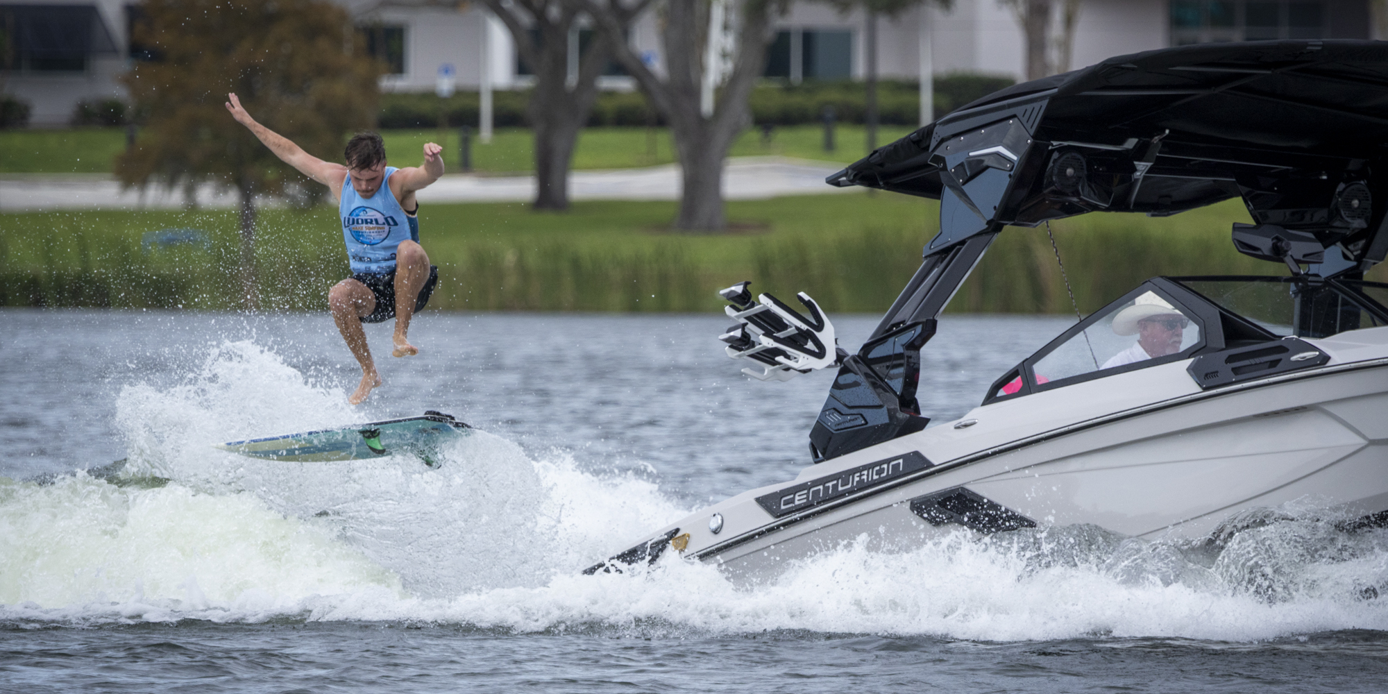 A person jumps off a wakeboard while riding behind a white motorboat on a lake, with trees and buildings visible in the background.