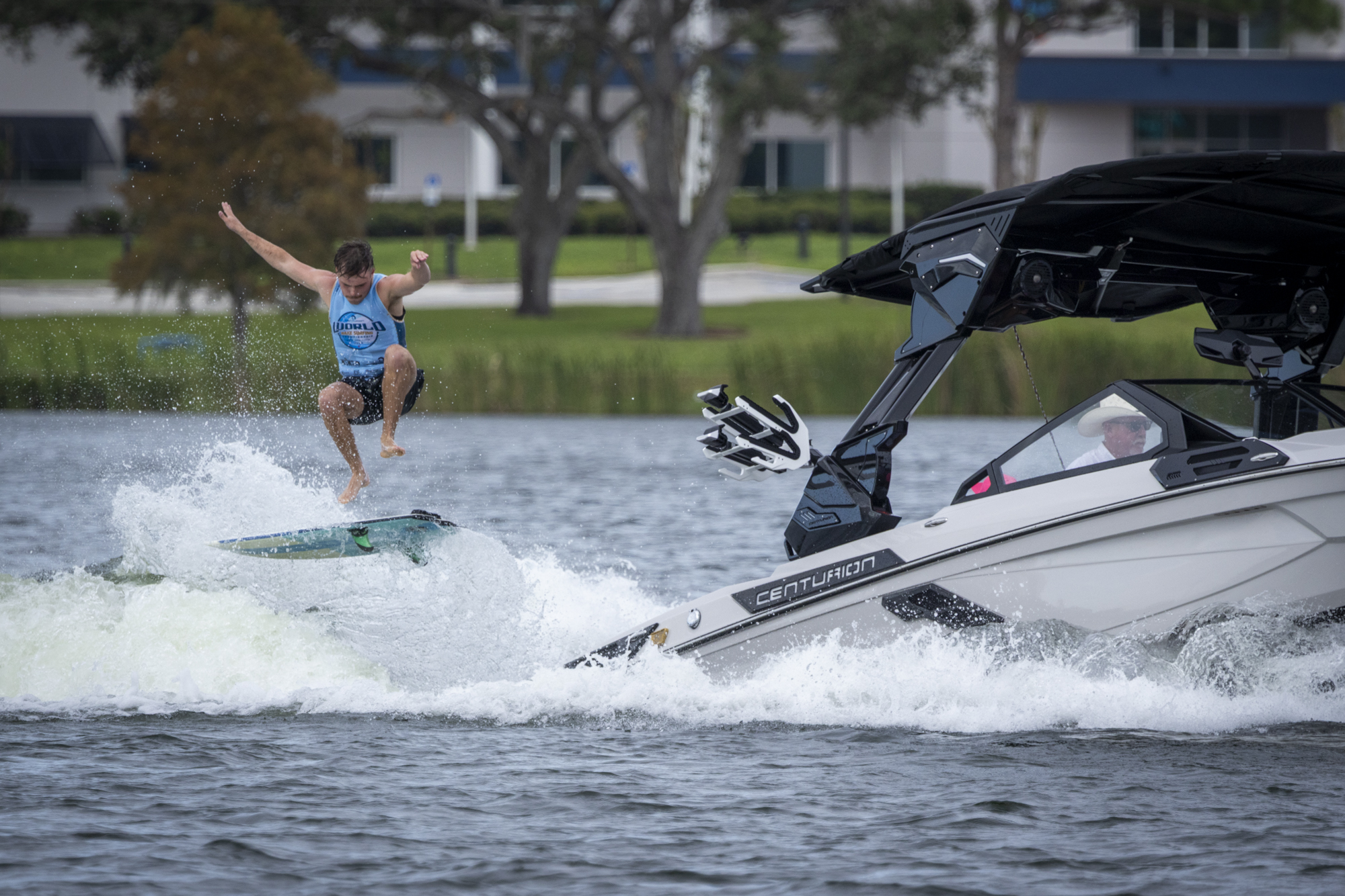 A person jumps off a wakeboard while riding behind a white motorboat on a lake, with trees and buildings visible in the background.