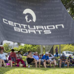 A group of people sit on chairs and blankets under a large "Centurion Boats" tent outdoors on a grassy area.