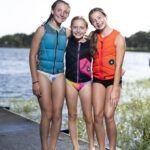 Three girls in swimsuits and life jackets stand on a dock by a lake, smiling at the camera with water and greenery in the background.
