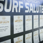 A display board with "SURF SALUTE" at the top features handwritten notes on white cards, each beginning with "I’m riding for," sharing personal motivations.