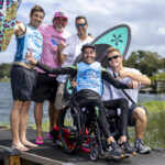 Five men, one in a wheelchair, pose and smile together on a dock by a lake, holding colorful paddleboards, with trees and water in the background.