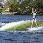 A person rides a wave on a pink and blue board with both arms raised, wearing a blue tank top and black shorts on a lake with trees and buildings in the background.