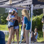 Several people stand and hug under a canopy labeled “Centurion Boats” at an outdoor event; others sit nearby, and a photographer films the scene.