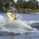 A person in a blue swimsuit rides a yellow wakesurf board on the water behind a boat, mid-jump, with trees and grass visible in the background.