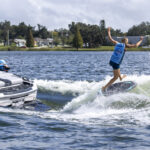 A person in a blue vest falls backwards off a wakesurf board behind a boat on a lake, while people watch from the boat.
