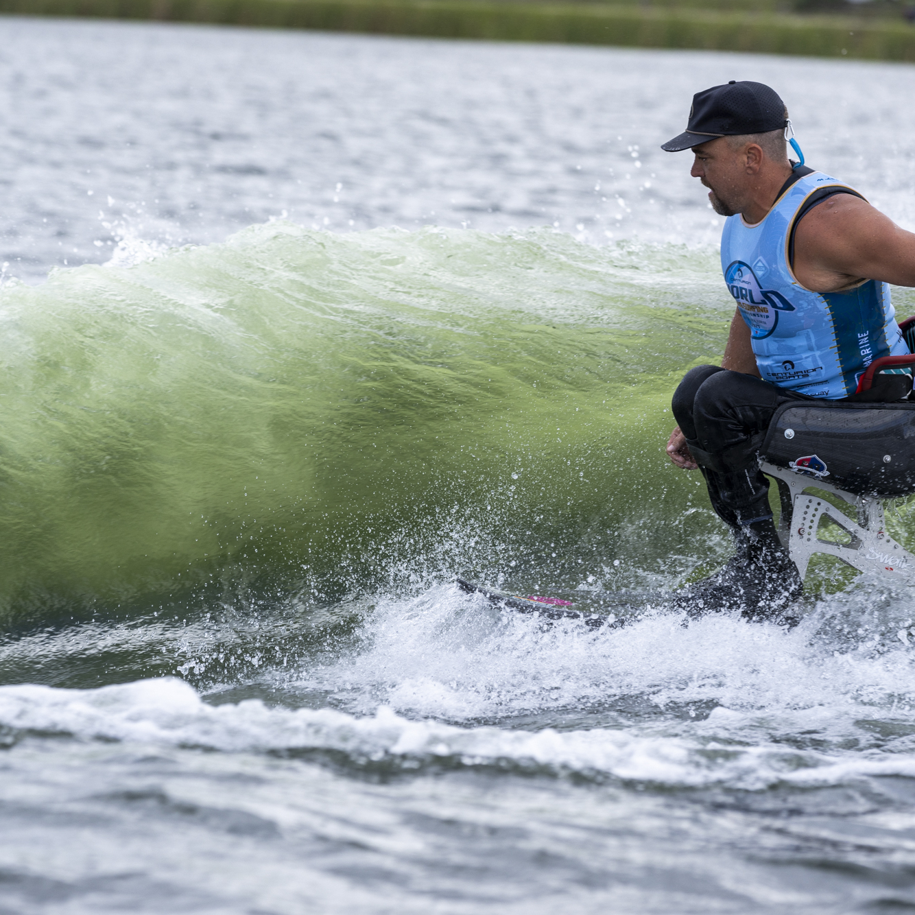 A man with a prosthetic leg is surfing on a wave, wearing a blue tank top, black cap, and black pants.