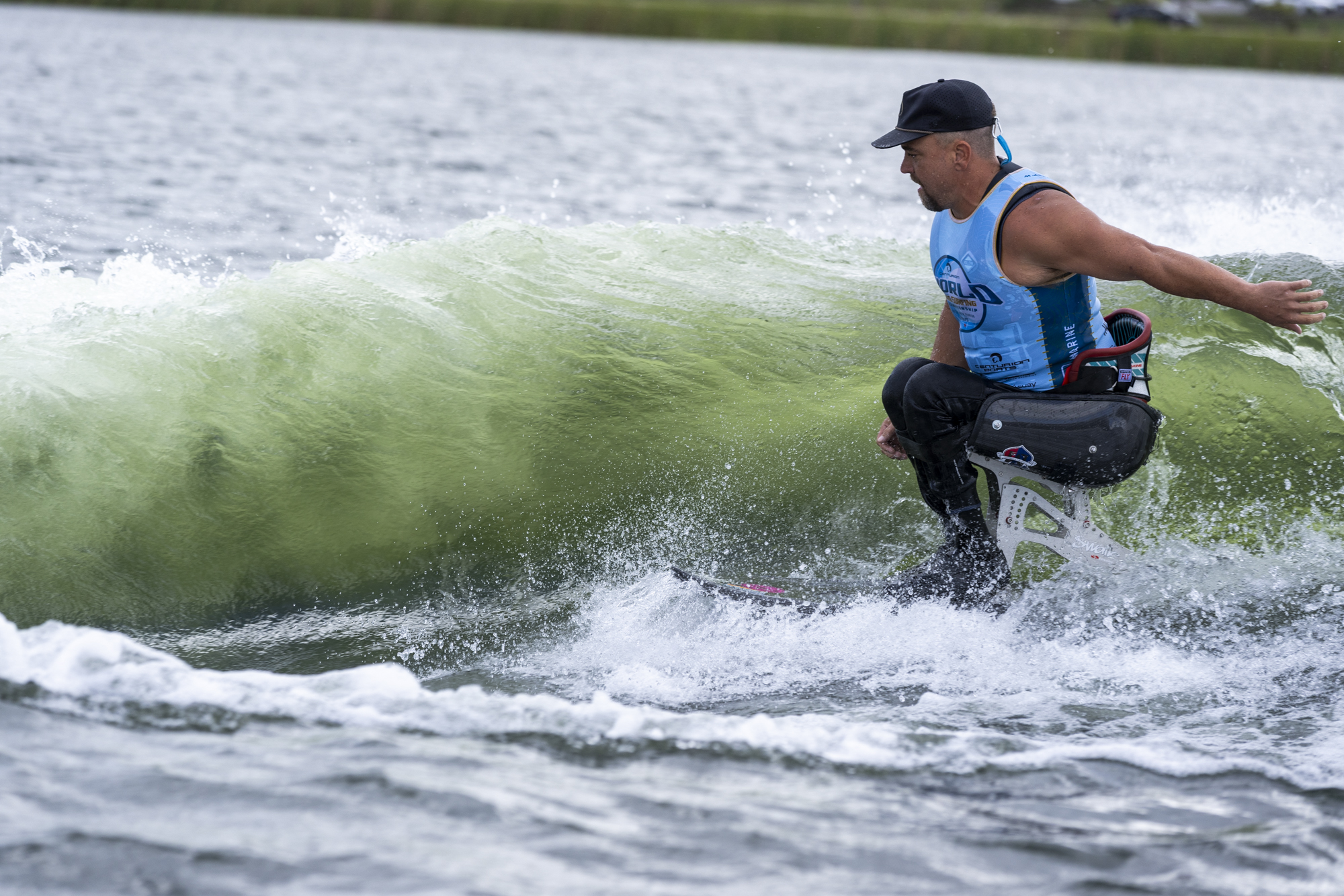 A man with a prosthetic leg is surfing on a wave, wearing a blue tank top, black cap, and black pants.