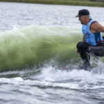 A man with a prosthetic leg wears a blue tank top and rides a wave on a surfboard in a body of water.