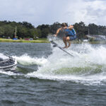 A person in a blue vest performs a jump trick on a wakeskate behind a motorboat on a lake, creating a spray of water.