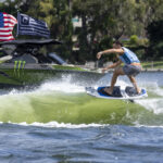 A person rides a wakesurf board on a wave created by a green boat with American flags and sponsor logos on a sunny day.