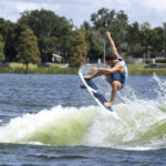 A person in a blue tank top and shorts performs a trick on a wakeboard, catching air above the water with trees and cars in the background.