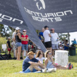 People sit and stand on a grassy area under a large Centurion Boats canopy at an outdoor event on a sunny day.