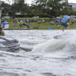 A person in blue shorts surfs behind a boat, performing a trick in mid-air, with spectators watching from a grassy shore in the background.