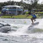 A person on a wakeboard performs a trick on a wave created by a boat, with spectators and a building visible in the background.