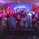 A group of people, some holding surfboards and trophies, gather on a stage under a tent with sponsor logos in the background; one person is in a wheelchair.