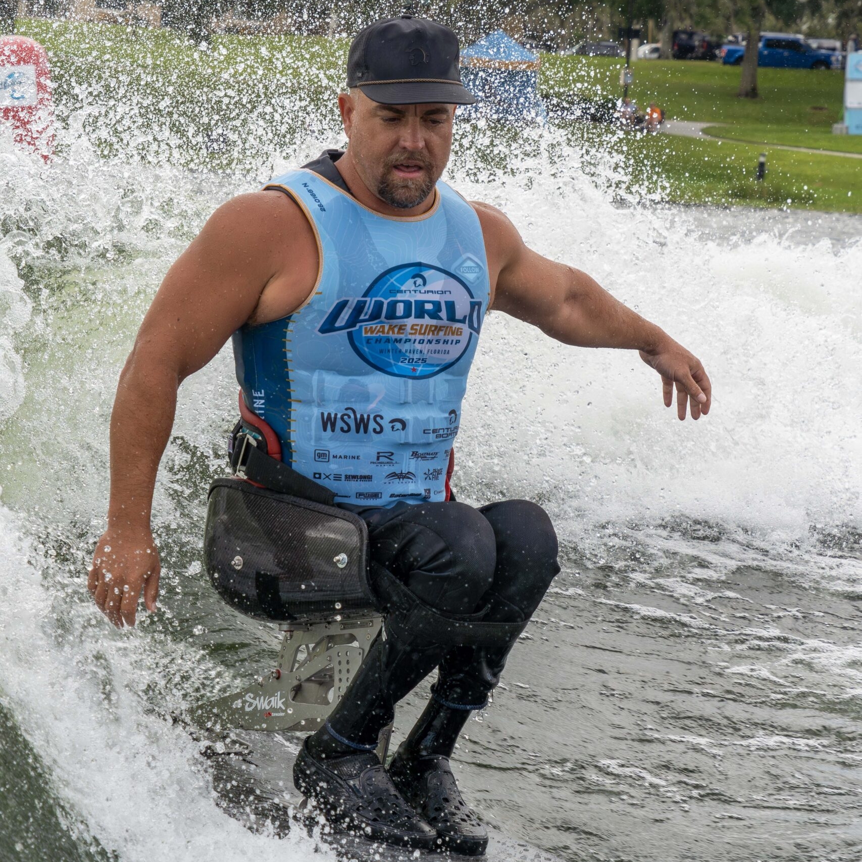 A man with a prosthetic leg is wakesurfing on a river, wearing a blue competition vest and a black cap, surrounded by splashing water.