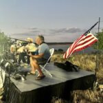 Three men play musical instruments on a small outdoor stage near a lake at sunset, with an American flag displayed to the right.