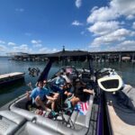 A group of people sit and pose for a photo on a docked speedboat under a sunny sky, with a marina and water in the background.