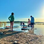 Four people stand and kneel near paddleboards at the edge of a calm body of water on a sunny day.