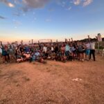 A large group of people pose together outdoors on a gravel area near water at sunset, many raising their arms and smiling at the camera.