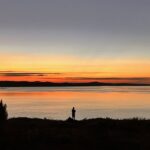 A person stands by a calm lake at sunset, silhouetted against an orange and blue sky with a bright star visible above the horizon.