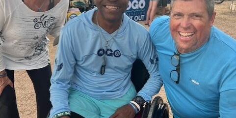 Three adults smile at an outdoor event booth; one is seated in a wheelchair, and two are standing on either side. They all wear casual, sporty clothing.