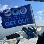 A blue flag reading "OGO Operation Get Out" is attached to a boat, with a wakeboard secured next to it over the water.