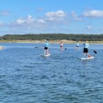 Four people are paddleboarding on a calm lake under a blue sky with scattered clouds, with trees visible in the background.