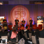 Four men stand and speak on stage at a conference, with attendees seated at tables. A 50th anniversary logo is displayed behind them along with banners and screens.