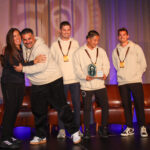 Five people stand smiling on a stage; one holds a plaque. Four wear matching light-colored hoodies and medals. The background features a brown curtain and leather chairs.