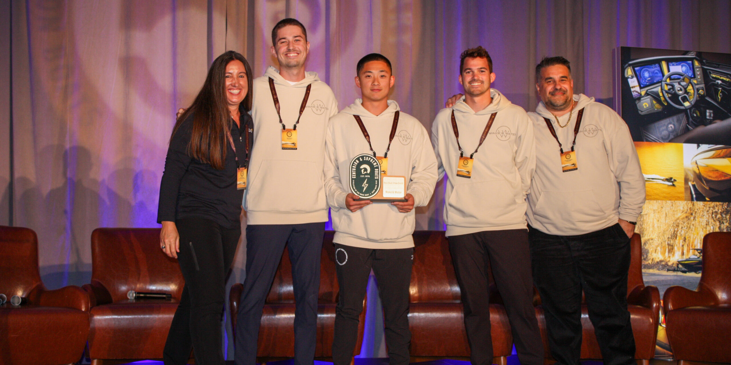 Five people stand on a stage, with one person in the center holding a plaque and small box. All are smiling and wearing event badges; three wear matching light-colored hoodies.