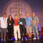 Six people stand on a stage, one holding a plaque. They are smiling and posing for a group photo in front of chairs and a backdrop with a large "50" projected on it.