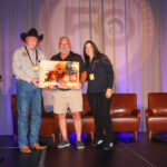 Three people stand on a stage holding a colorful award plaque, with empty chairs and a podium nearby and a large logo projected in the background.
