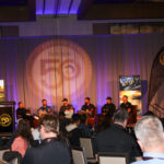 A panel of six people speaks on stage at a conference in front of an audience, with banners and screens displaying agricultural images and a "50" logo in the background.