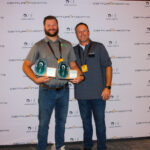 Two men stand in front of a Centurion Boats backdrop; one holds two award plaques, both smiling and wearing conference badges.