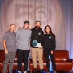 Four people stand on a stage in front of chairs and a curtain; one person holds an award plaque, and they all smile at the camera.