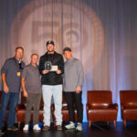 Four men stand on a stage, with one holding an award plaque. Three brown chairs and a display with boating images are visible in the background.