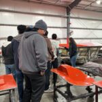 A group of people stands in a workshop near several red-painted parts and a boat hull, with industrial shelves and equipment in the background.